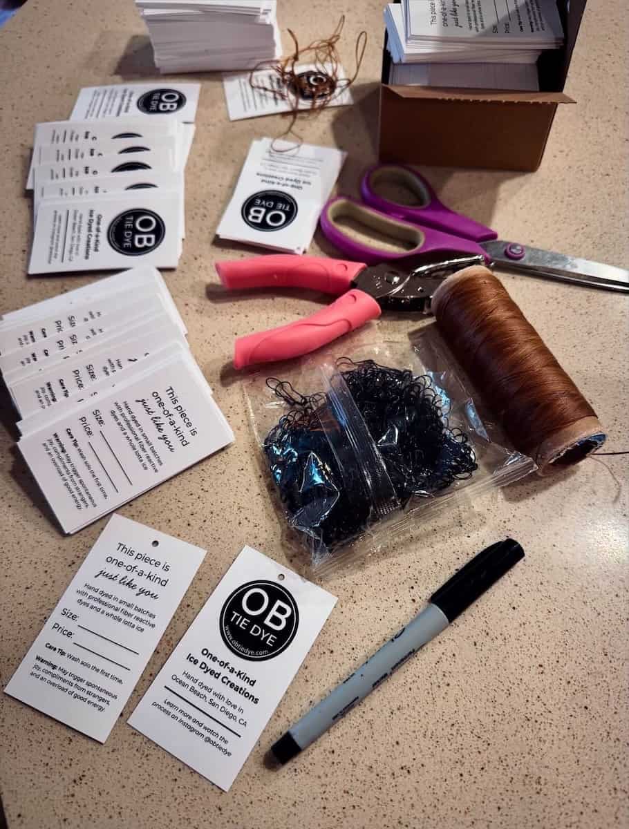 Clothing finishing supplies including scissors, thread, and OB Tie Dye hang tags on a table.