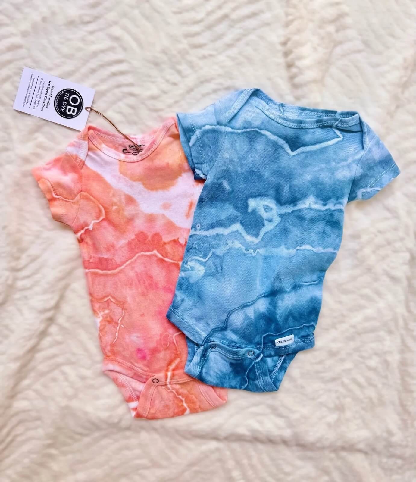Two tie-dye baby onesies, one pink and one blue, on a textured surface with a brand tag.