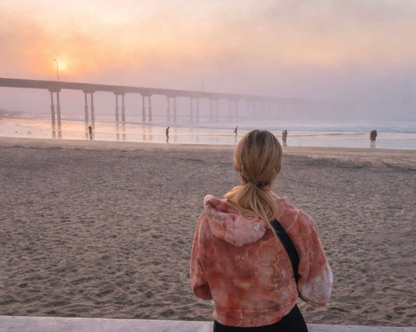 OB Tie Dye founder Jamie Peterson wearing a premium ice dyed cropped hoodie in Sunset Cliffs colorway on Ocean Beach at sunset, facing the OB pier in San Diego. 
