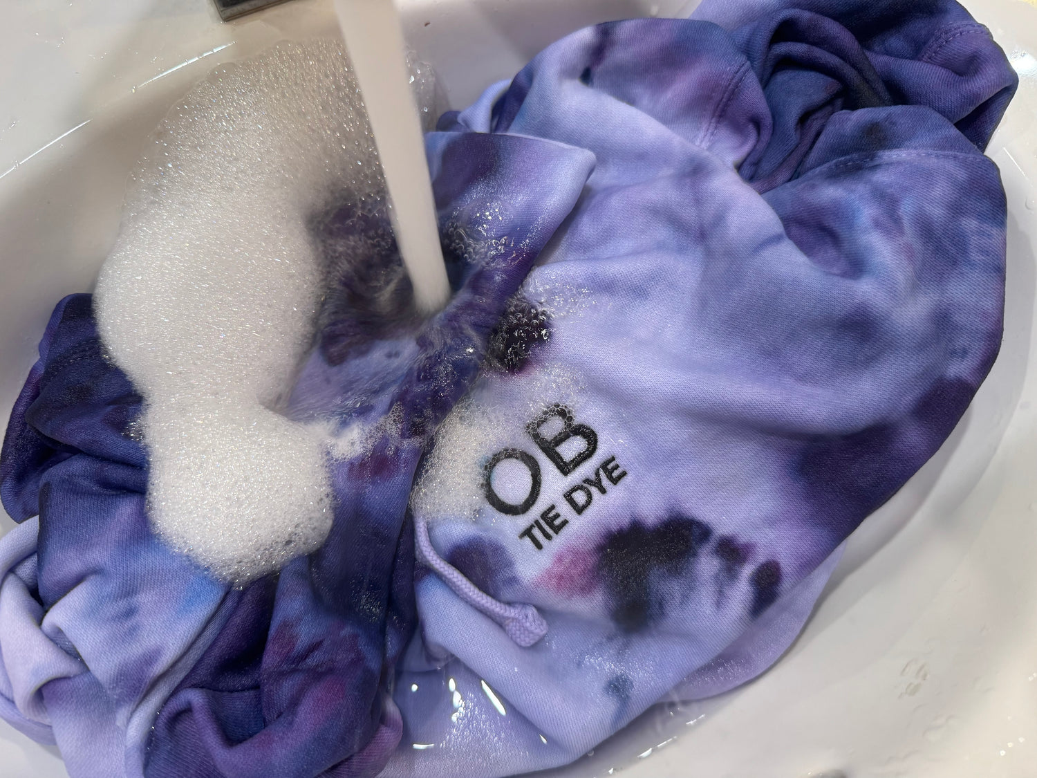 An image of a custom embroidered OB TIE DYE hoodie in shades of purple being washed out in the sink to remove the dye.
