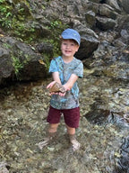 Child holding a stone in a stream with rocks and water while wearing a hand ice dyed kids tee with green and blue patterns  from OB Tie Dye.